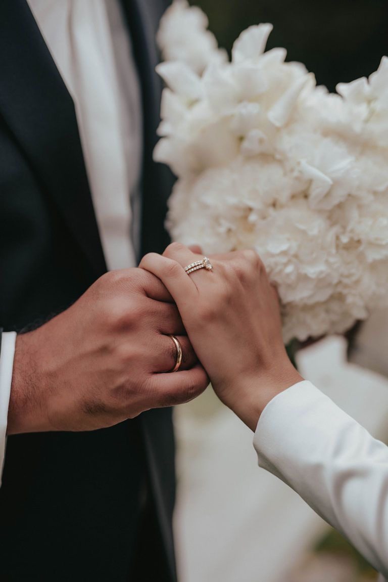 Mains des mariés avec leurs alliances devant un bouquet blanc